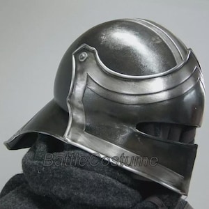 K&ouml;nnte beinhalten: Dunkelgrauer mittelalterlicher Helm mit silbernen Akzenten. Der Helm hat ein Visier und eine abgerundete Oberseite mit einem Mittelgrat. Der Text "Battle Costume" ist auf dem unteren Teil des Helms sichtbar.