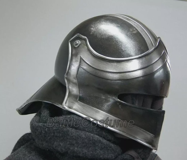 K&ouml;nnte beinhalten: Dunkelgrauer mittelalterlicher Helm mit silbernen Akzenten. Der Helm hat ein Visier und eine abgerundete Oberseite mit einem Mittelgrat. Der Text "Battle Costume" ist auf dem unteren Teil des Helms sichtbar.