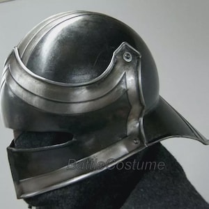 K&ouml;nnte beinhalten: Dunkelgrauer Metallhelm mit Visier und silberner Zierleiste. Der Helm hat eine abgerundete Oberseite mit vertikalen Rippen und einem gebogenen Stirnband. Der Text "BattleCostume" ist auf der Seite sichtbar.