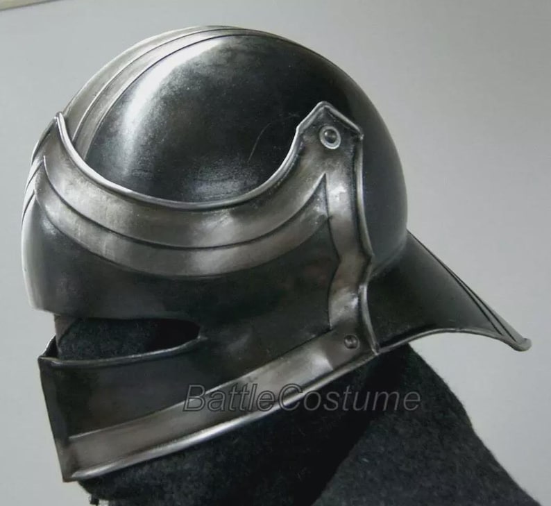 K&ouml;nnte beinhalten: Dunkelgrauer Metallhelm mit Visier und silberner Zierleiste. Der Helm hat eine abgerundete Oberseite mit vertikalen Rippen und einem gebogenen Stirnband. Der Text "BattleCostume" ist auf der Seite sichtbar.