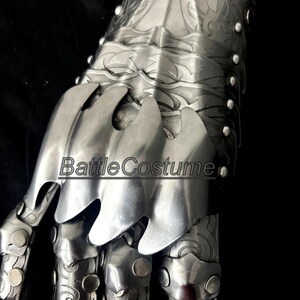 May include: Close-up of a silver metal gauntlet with articulated fingers and intricate engraved designs. The gauntlet features overlapping plates and rivets, with the text "BattleCostume" visible.