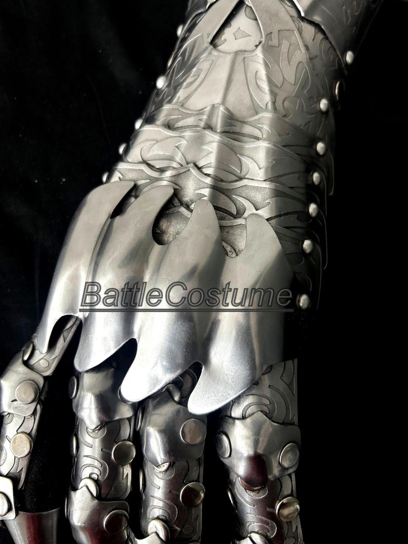 May include: Close-up of a silver metal gauntlet with articulated fingers and intricate engraved designs. The gauntlet features overlapping plates and rivets, with the text "BattleCostume" visible.