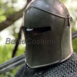 May include: A dark gray medieval helmet with a visor and the text "Battle Costume". The helmet is made of metal and has a smooth surface. The helmet is part of a medieval costume.