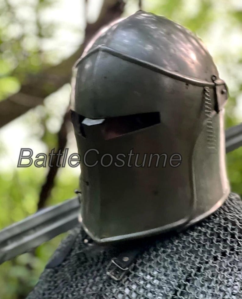 May include: A dark gray medieval helmet with a visor and the text "Battle Costume". The helmet is made of metal and has a smooth surface. The helmet is part of a medieval costume.