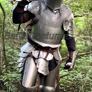 May include: A full suit of medieval armor, including a helmet, chainmail, and plate armor, with a sword resting on the shoulder. The armor is silver-colored and worn in a forest setting. The text "Battle Costume" is visible.