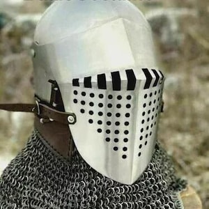 May include: A silver medieval helmet with a perforated visor. The helmet is part of a chainmail coif and has a leather strap. The text "BattleCostume" is visible at the top. The helmet is a historical item.
