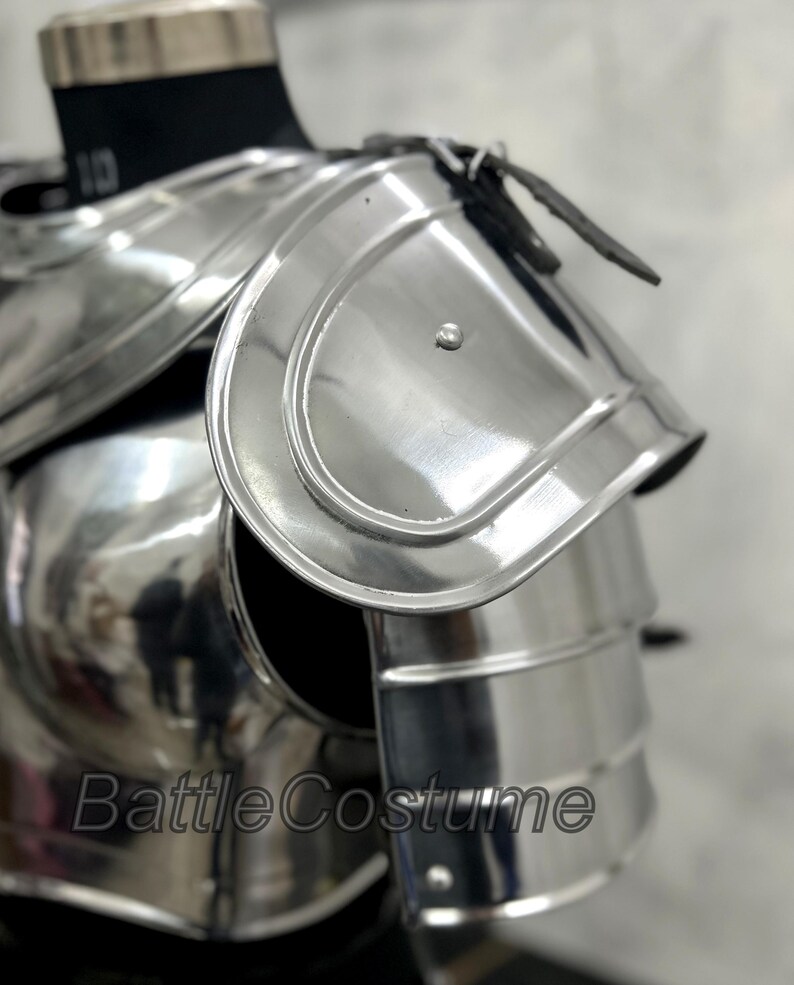 May include: Polished silver shoulder armor with layered plates and a curved design. The armor is displayed on a black mannequin and features a leather strap. The text "BattleCostume" is visible on the armor.