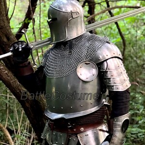 May include: A full suit of medieval armor, including a helmet, chainmail, and plate armor, is displayed. The silver-colored armor is worn with black gloves and a sword. The image is set in a forest, with the text "BattleCostume" visible.