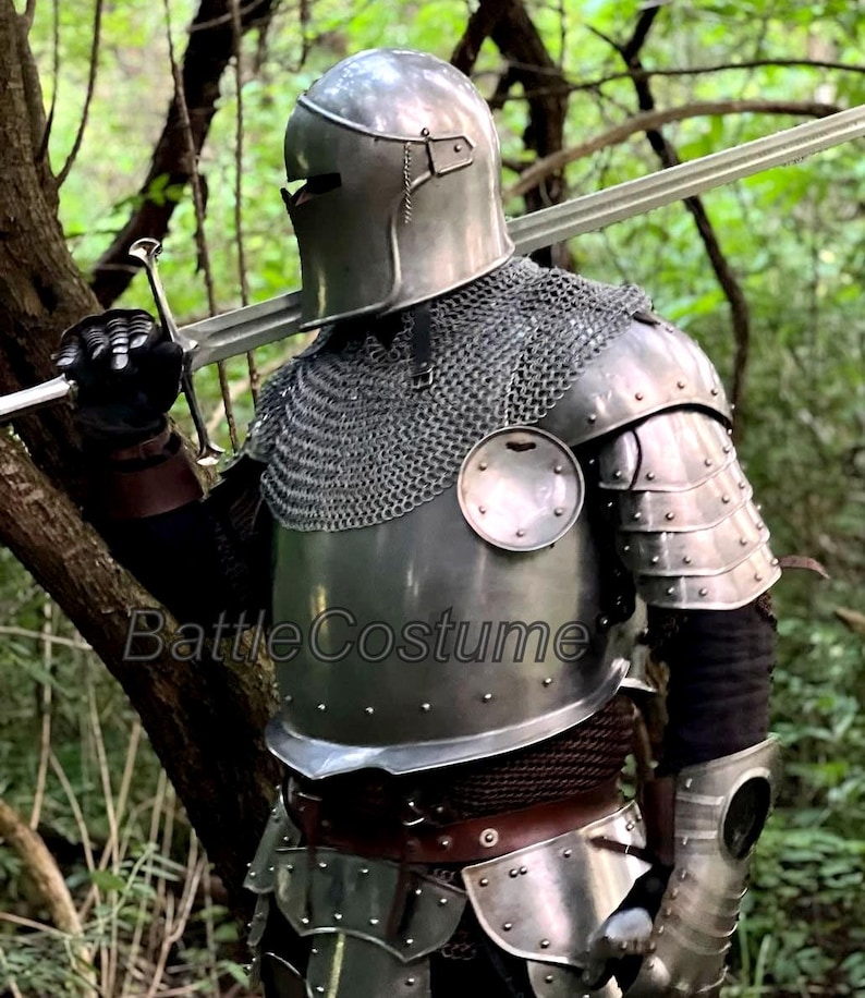 May include: A full suit of medieval armor, including a helmet, chainmail, and plate armor, is displayed. The silver-colored armor is worn with black gloves and a sword. The image is set in a forest, with the text "BattleCostume" visible.