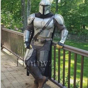 May include: A silver and gray Mandalorian costume, complete with helmet, chest armor, and leg guards. The costume includes a black cape and utility belt. The person is leaning against a wooden railing, with a green background.