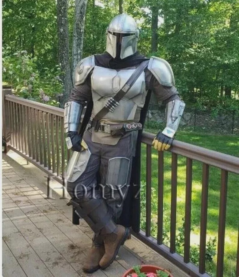 May include: A silver and gray Mandalorian costume, complete with helmet, chest armor, and leg guards. The costume includes a black cape and utility belt. The person is leaning against a wooden railing, with a green background.