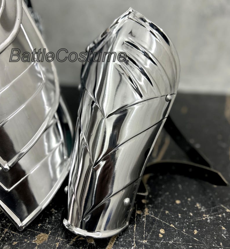 May include: Highly polished silver metal arm armor with riveted accents. The armor has a reflective surface and is designed with a series of angled plates. The text "BattleCostume" is visible on the armor.
