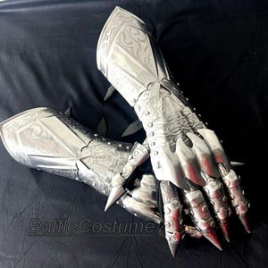 May include: A pair of silver-colored metal gauntlets with intricate engraved designs. The gauntlets feature articulated fingers and sharp, pointed spikes. The text "BattleCostume" is visible in the image.