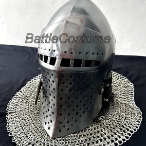 Puede incluir: Un yelmo de caballero de acero pulido con una parte superior puntiaguda y una protección facial perforada. El yelmo descansa sobre una cofia de cota de malla circular. El texto "Battle Costume" es visible en el yelmo.