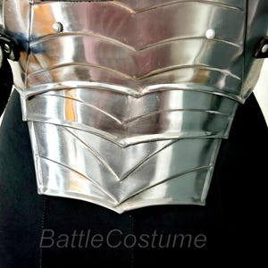May include: Close-up of a polished, silver metal chest armour. The armour features layered, chevron-shaped plates and is secured with black leather straps. The text "Battle Costume" is visible at the bottom.