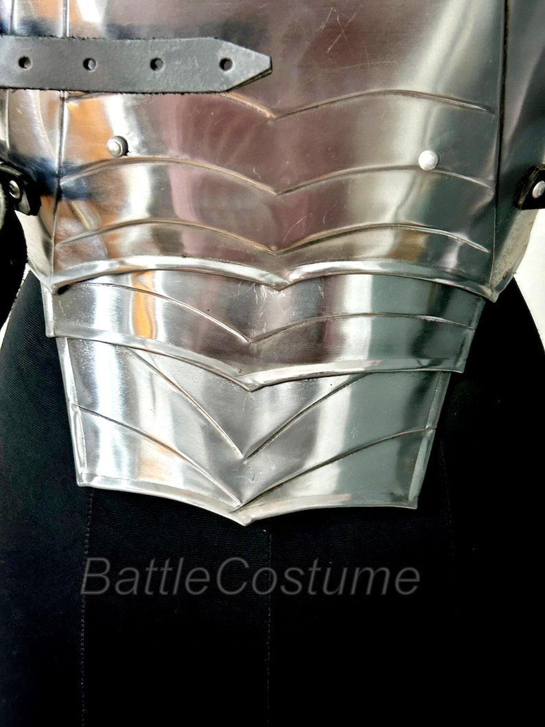May include: Close-up of a polished, silver metal chest armour. The armour features layered, chevron-shaped plates and is secured with black leather straps. The text "Battle Costume" is visible at the bottom.