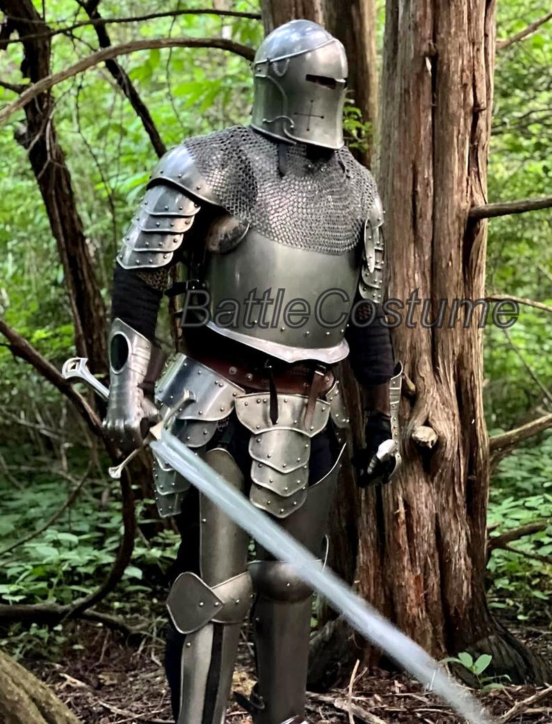 May include: A full suit of medieval armor, including a helmet, chainmail, and plate armor, with a long sword. The armor is silver-colored and worn by a person standing in a forest. The text "Battle Costume" is visible.