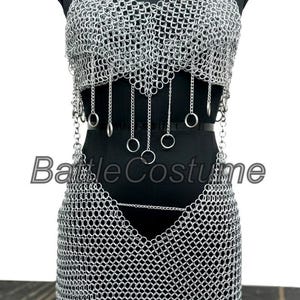 Puede incluir: Un conjunto de disfraz de cota de malla plateada, que incluye un top halter y una falda, exhibido en un maniqu&iacute; negro. La parte superior presenta anillos colgantes y la falda tiene un dobladillo puntiagudo. El texto "Battle Costume" es visible.