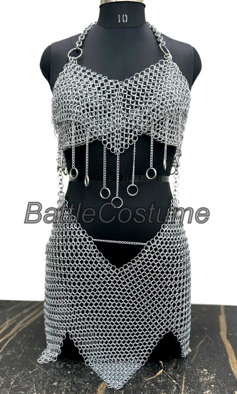 Puede incluir: Un conjunto de disfraz de cota de malla plateada, que incluye un top halter y una falda, exhibido en un maniqu&iacute; negro. La parte superior presenta anillos colgantes y la falda tiene un dobladillo puntiagudo. El texto "Battle Costume" es visible.