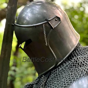 May include: A close-up of a medieval steel helmet with a visor and chainmail. The helmet is a dark gray color and has a smooth, reflective surface. The text "BattleCostume" is visible on the helmet.