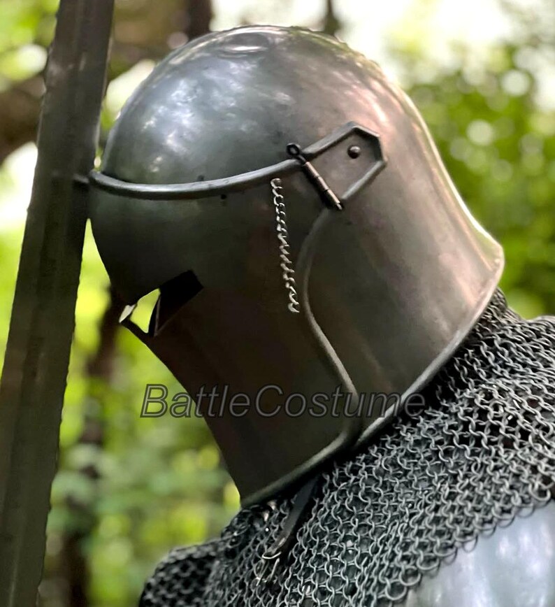 May include: A close-up of a medieval steel helmet with a visor and chainmail. The helmet is a dark gray color and has a smooth, reflective surface. The text "BattleCostume" is visible on the helmet.