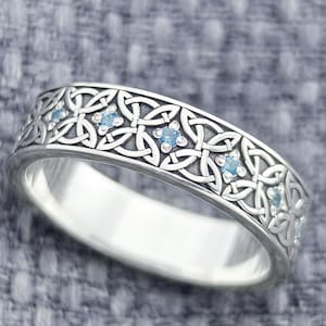 May include: Silver ring with a Celtic knot design and small blue gemstones. The ring has an intricate pattern of interwoven knots around the band. The ring is on a gray textured surface. The logo "JEWELRY ETERNITY" is at the bottom.
