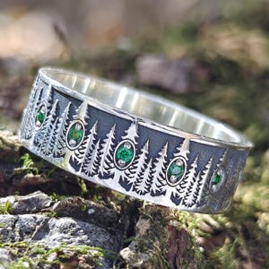 Peut inclure: Bague en argent avec un motif de forêt. La bague présente une bande avec des arbres gravés et des pierres précieuses vertes de forme ovale. La bague est posée sur un fond naturel de mousse et de bois.