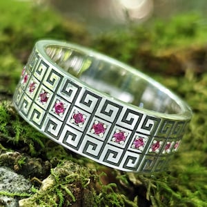 May include: A silver ring with a Greek key design and small red gemstones. The ring is set against a backdrop of green moss and wood, with the Jewelry Eternity logo visible in the lower left corner.