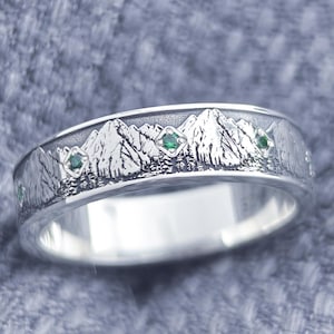 May include: A silver ring with a mountain range design. The ring features a detailed engraving of snow-capped mountains and small green gemstones. The ring is set against a textured, light blue background. The ring is a piece of jewelry.