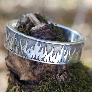 Può includere: Un anello d'argento con un design a fiamma. L'anello presenta una fascia con un motivo ripetuto di fiamme stilizzate. L'anello è esposto su un pezzo di legno ricoperto di muschio verde, con il logo Jewelry Eternity visibile.
