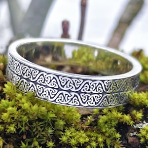 May include: A silver ring with an intricate Celtic knot pattern. The ring is displayed on a moss-covered log, with the Jewelry Eternity logo visible. The ring's design features repeating geometric shapes, creating a textured appearance.