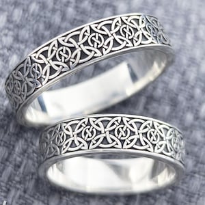 Könnte beinhalten: Zwei silberne Ringe mit einem keltischen Knotenmuster. Die Ringe werden auf einer grauen, strukturierten Oberfläche präsentiert. Die Ringe haben ein detailliertes Muster um das Band. Die Ringe sind von Jewelry Eternity.
