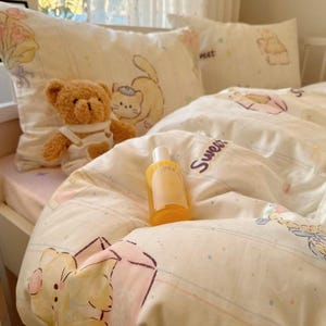 May include: A cream-coloured bed set with a teddy bear, pillows, and a bottle of lotion. The bedding features cartoon illustrations. The duvet cover and pillowcases have a soft texture. The word "Sweet" is printed on the duvet.
