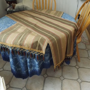 May include: A striped table runner with a gold and brown trim and brown tassels. The runner is on a blue table with a floral pattern.