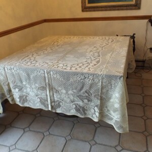 May include: A white lace tablecloth with a floral pattern covers a square table. The table is in a room with beige tile flooring.