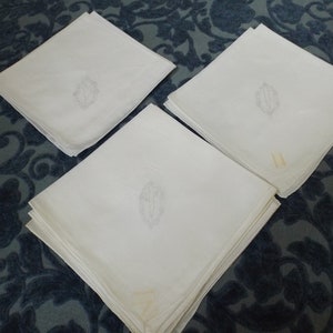 May include: Three white linen napkins with embroidered initials in the center. The napkins are folded and stacked on a blue patterned surface.
