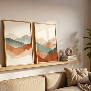 May include: Two framed landscape art prints with abstract mountain designs in shades of rust, teal, and beige. The prints are displayed on a wooden shelf above a beige sofa, creating a warm and inviting atmosphere.