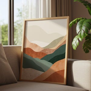 Op de afbeelding: Een ingelijste kunstdruk met een abstract landschap met gelaagde heuvels in tinten beige, roest en groen. Het kunstwerk wordt weergegeven op een beige bank, met een grote kamerplant op de achtergrond. Het frame is gemaakt van lichtgekleurd hout.