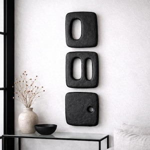 May include: Three rectangular black wall art pieces with textured surfaces arranged vertically. The top piece has one large oval cutout, the middle piece has two vertical oval cutouts, and the bottom piece features a single small circular hole.