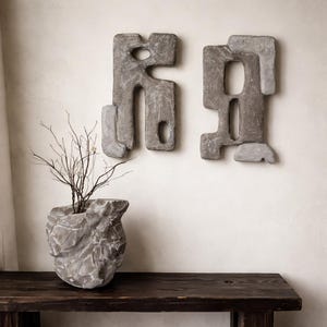 May include: Two grey, abstract wall sculptures with irregular shapes hang on a light-coloured wall. A textured, grey stone-like planter holds dried branches, sitting on a dark wooden table. The sculptures and planter create a modern, minimalist aesthetic.