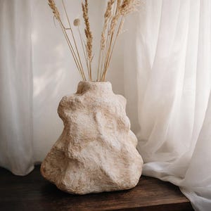 May include: A beige, textured vase with an organic, rock-like shape. The vase holds dried, tan-coloured grasses, creating a natural, minimalist aesthetic. The vase is on a dark wooden surface, with a white curtain in the background.