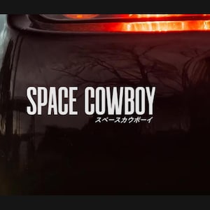May include: A black car bumper featuring a white decal with the words "SPACE COWBOY" in capital letters. Beneath the English text is Japanese text. The car's taillight is visible at the top, glowing red.
