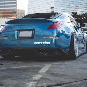 May include: A blue sports car with a spoiler and the text "DRIFT KING" in white. The car has a black rear diffuser and dual exhaust pipes. The car is parked on an asphalt surface.