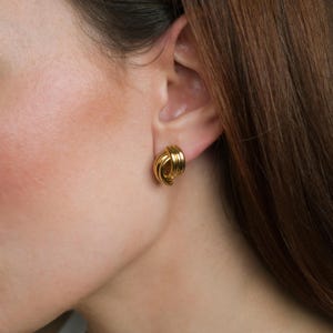 Peut inclure: Gros plan d'une boucle d'oreille dorée. La boucle d'oreille est de style clou avec un design noué. La boucle d'oreille est faite d'un matériau métallique brillant. La boucle d'oreille est sur l'oreille d'une personne aux cheveux bruns.