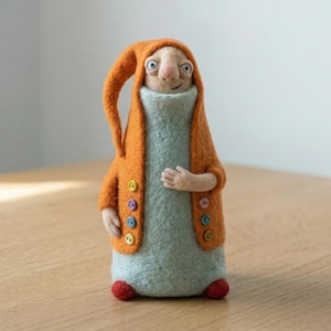May include: A whimsical felted figure with an orange hooded jacket and a light blue dress. The jacket has colorful buttons. The figure has a smiling face and red feet. The handmade doll is standing on a wooden surface.
