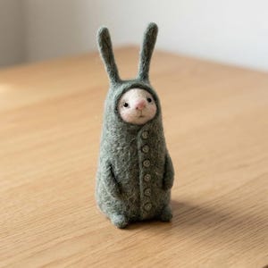 Puede incluir: Figura de conejo de fieltro hecha a mano, con un abrigo con capucha y botones. El conejo tiene orejas largas y una nariz rosa. El abrigo es de color verde apagado y la figura está sobre una superficie de madera.