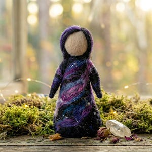 May include: A handmade felt doll with a cream-colored head and a dark purple hood. The doll's dress is a blend of dark blue, purple, and pink, resembling a galaxy. The doll is standing on a wooden surface with moss and a clear crystal.