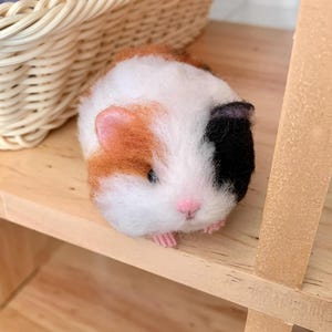 May include: A handmade guinea pig figurine with a white, brown, and black coat, pink nose, and tiny pink feet. The felted animal is positioned on a wooden shelf, with a wicker basket partially visible in the background.
