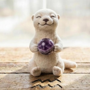 May include: A handcrafted, smiling otter figurine made of white felt, holding a purple crystal ball. The otter has closed eyes and a happy expression. The figurine is sitting on a wooden surface.