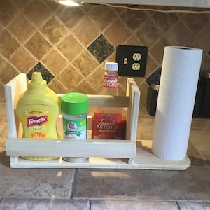 Pode incluir: Um organizador de cozinha de madeira clara com uma garrafa de mostarda amarela, um frasco de especiarias com tampa verde e um pacote de ketchup. Um rolo de papel toalha está à direita. O organizador está em uma bancada.